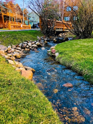 A clear stream with rocks and grass, surrounded by wooden structures and trees.