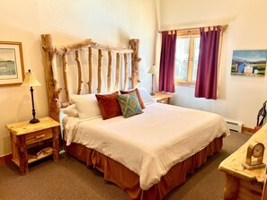 A bedroom with a wooden headboard, a bed with white bedding, a nightstand with a lamp, a window with curtains, and a painting on the wall.