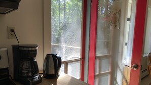 Et køkken med kaffemaskine og elkedel på en træbordplade, et vindue med udsigt til træer, og en rød dør.