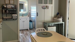 A kitchen with a white cabinet, a sink, a hanging rack with jars, and a table with a placemat. - Tiny for Two + Small Dog OK! -  Near BEACH, SPORTS COMPLEX, OWA, TANGER (Foley)