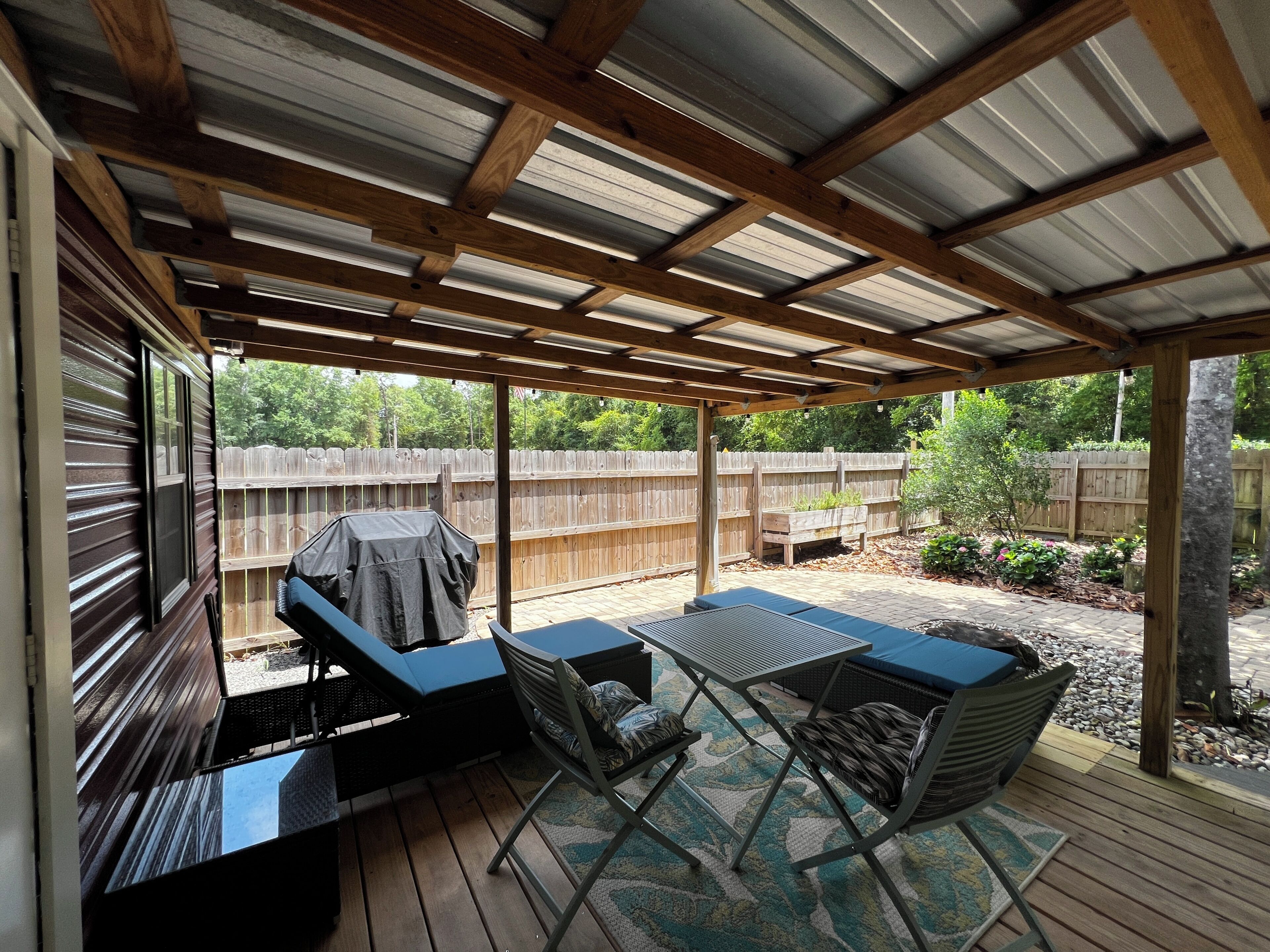 A covered patio area with a grill, seating, and a view of a wooden fence and garden.