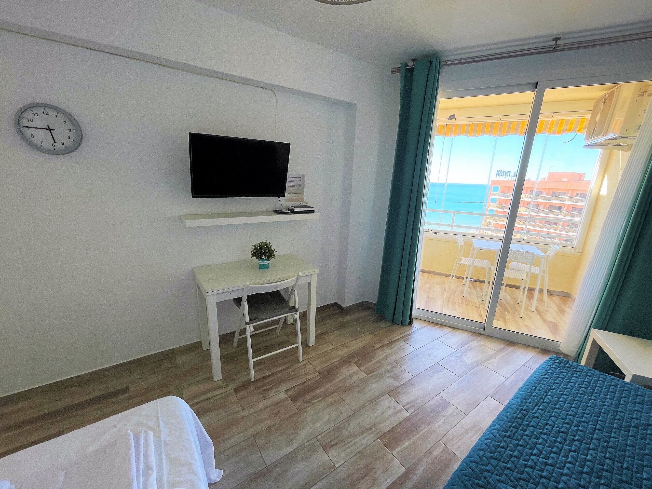 A room with a bed, a desk with a chair, a television, and a view of the sea through a sliding door.