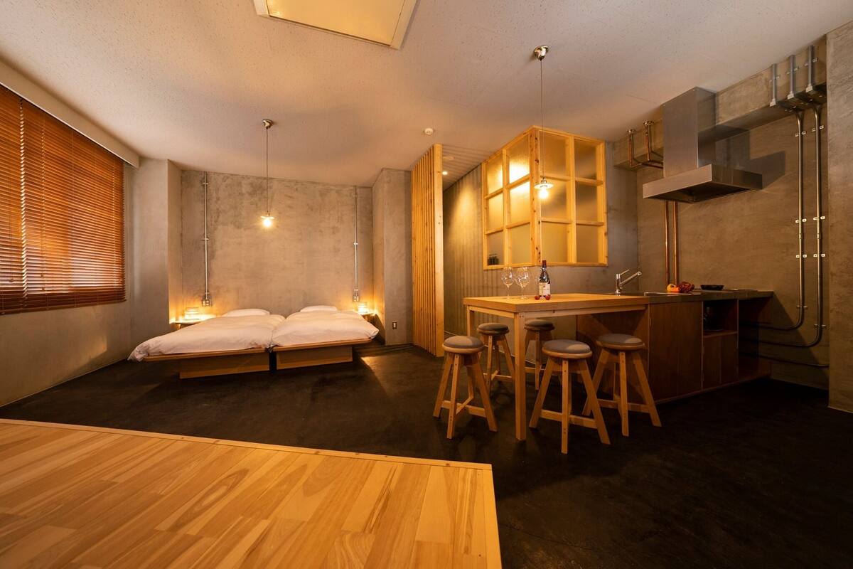Una habitación compacta con cama, un pequeño rincón de cocina con taburetes y una mesa de comedor.