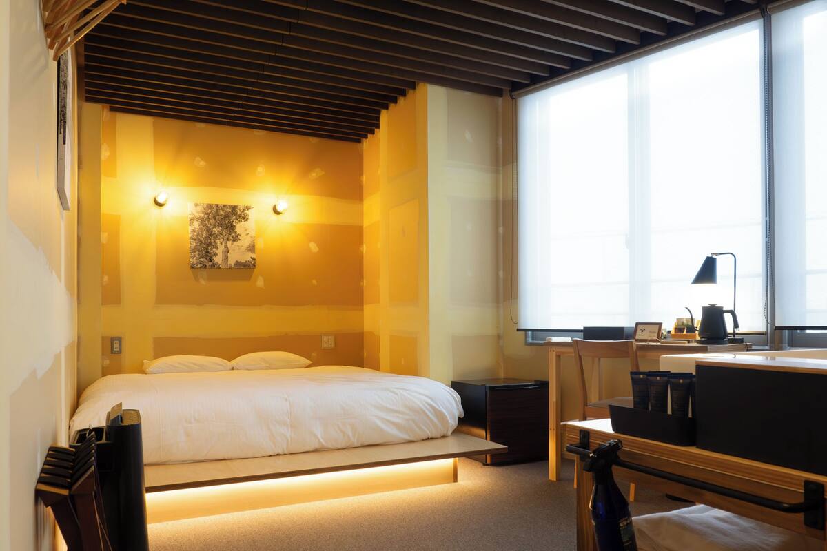 Una habitación de hotel moderna con cama, mesita de noche, lámpara y un ventanal grande.