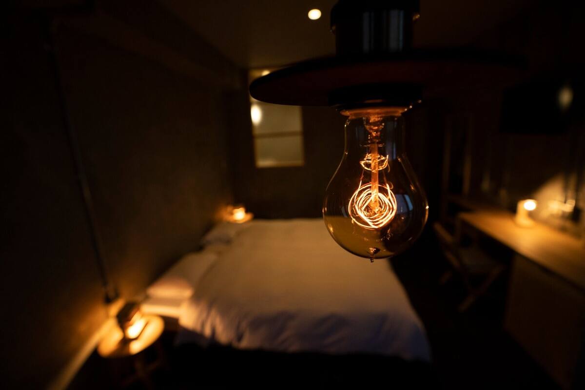 Una habitación tenuemente iluminada con una bombilla colgante, una cama y una mesita de noche con lámpara.