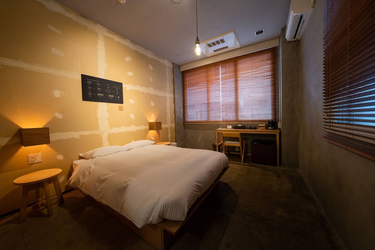 Un dormitorio con cama, mesitas de noche, un escritorio y una ventana con persianas.