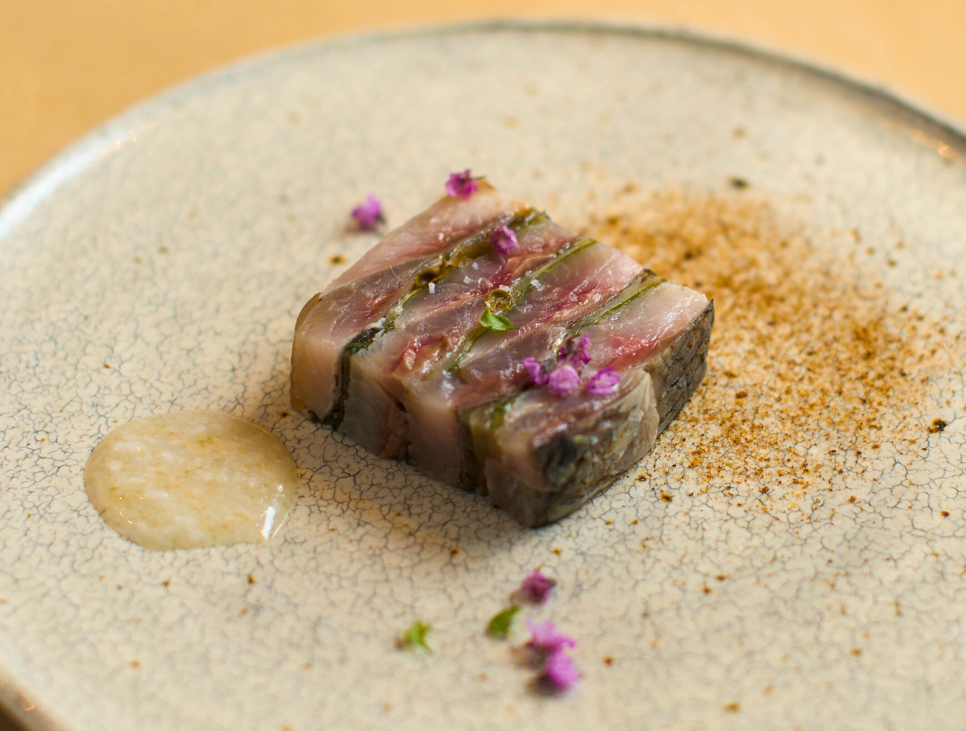 Un plato con carne en lonchas finas, adornado con hierbas y flores sobre una vajilla de cerámica texturizada.