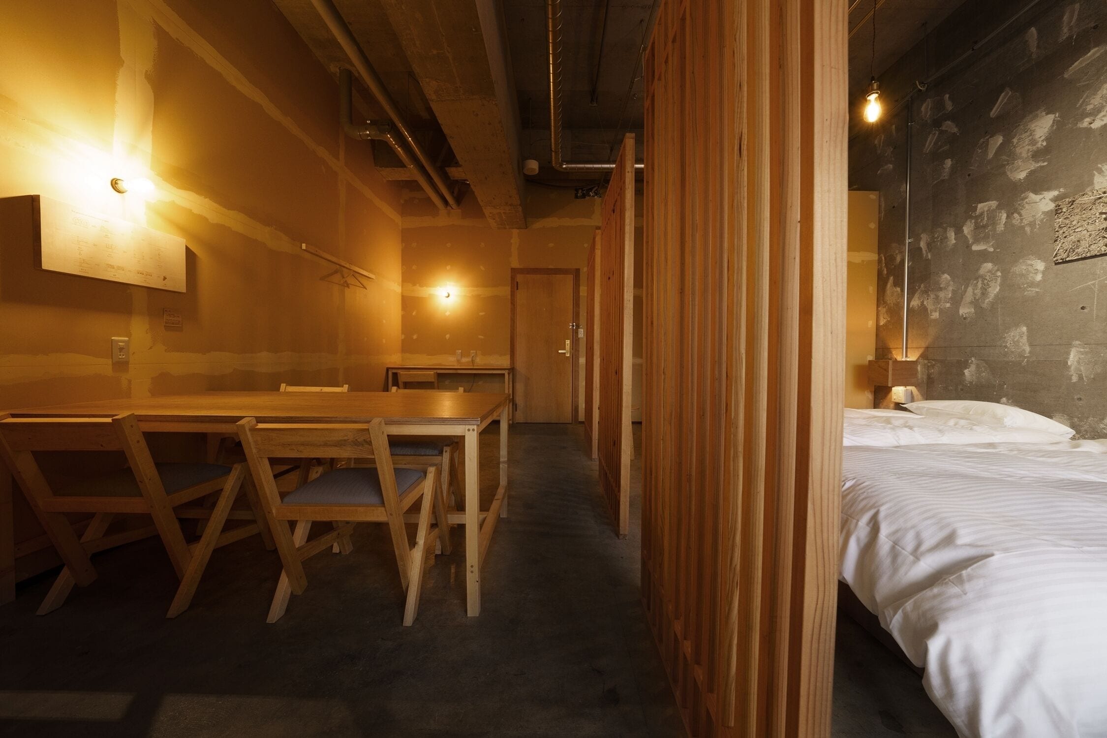 A hotel room with a wooden dining table and chairs, a bed with white bedding, and a wall with exposed brick.