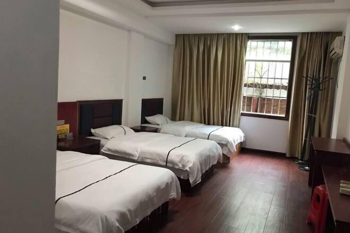 Une chambre d’hôtel avec trois lits simples, un plancher en bois, une fenêtre avec des rideaux et un bureau avec une chaise.