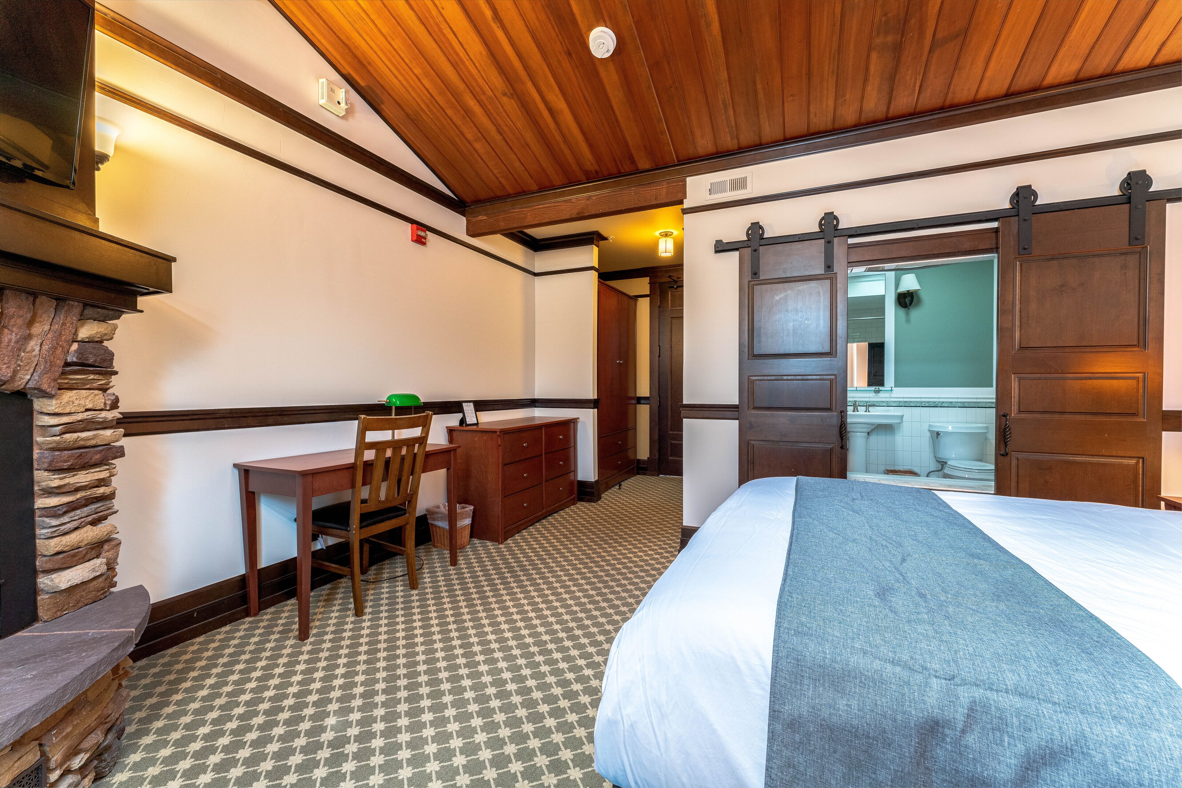 A hotel room with a bed, a desk, a chair, a stone accent wall, and a wooden ceiling.