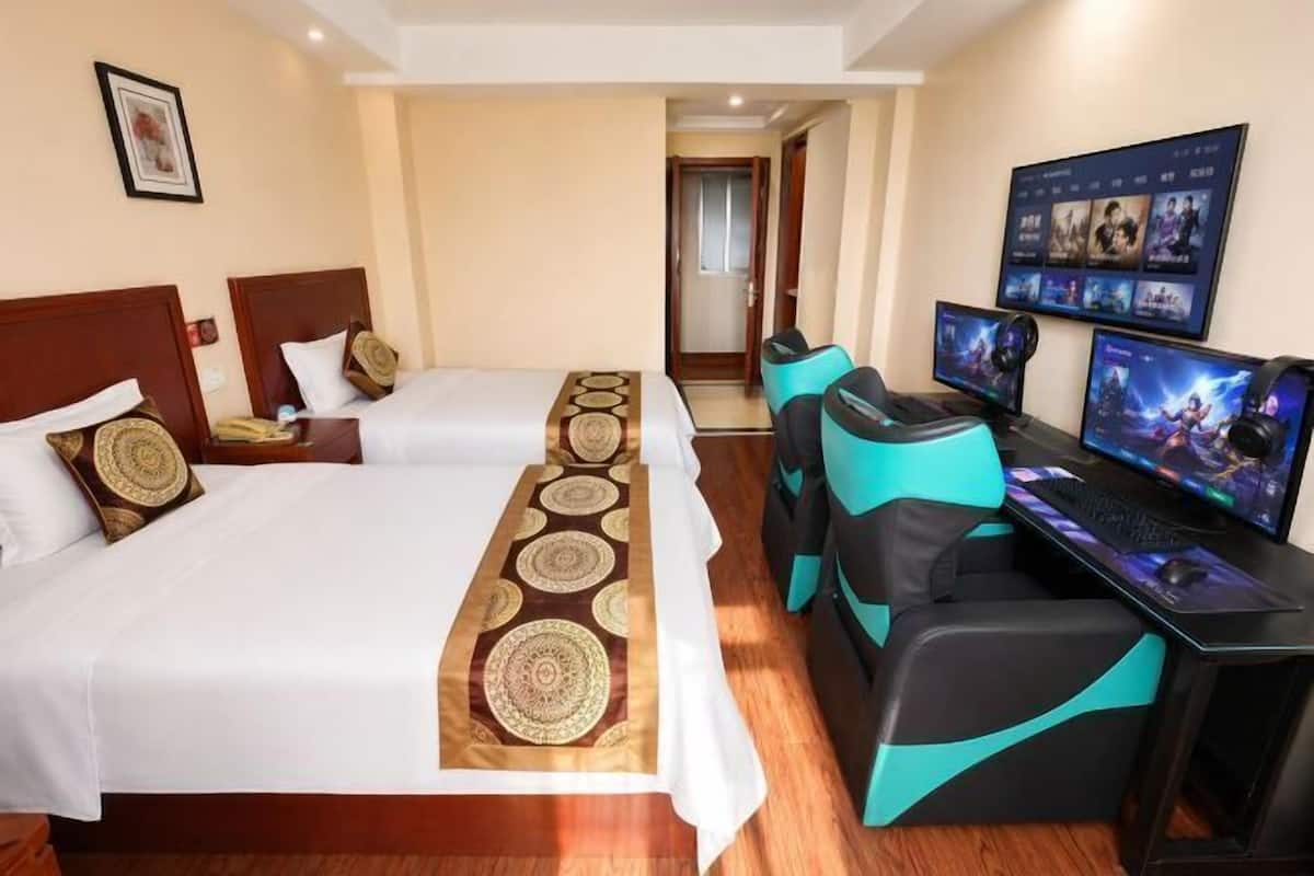 A hotel room with two beds, a desk with a computer, and a TV displaying a game.