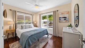 Une chambre à coucher comprenant un lit, des tables de chevet, un ventilateur de plafond, une fenêtre avec des rideaux et une vue sur la verdure à l’extérieur.