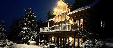 A snow-covered wooden lodge with a lit balcony and a staircase leading to the entrance.