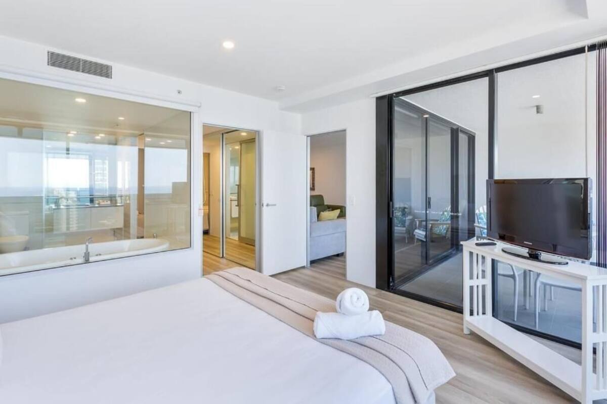 A modern hotel room with a large bed, a TV, a balcony with a glass door, and a view of the city.
