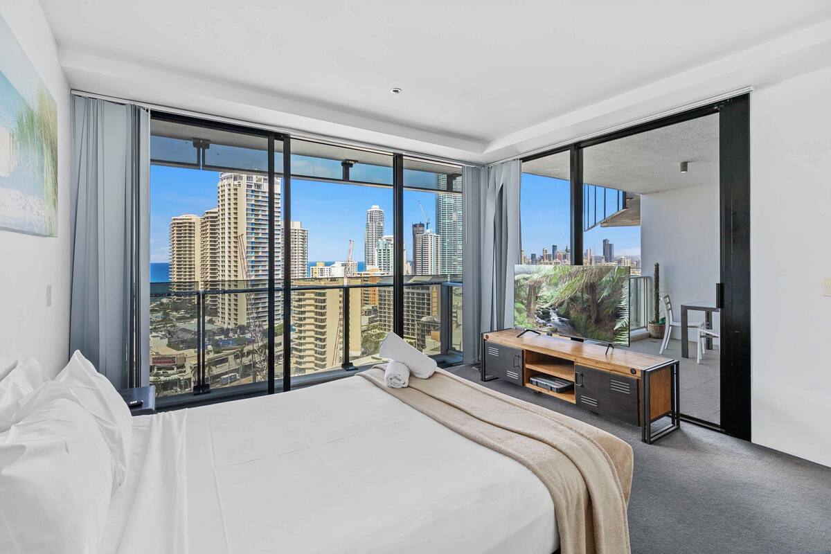A modern hotel room with a large bed, a wooden desk, and a view of high-rise buildings.