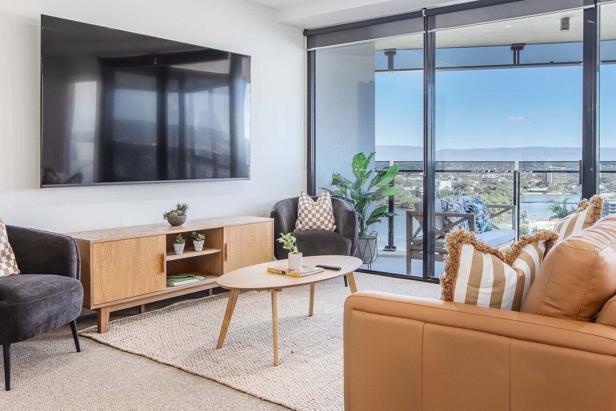 A modern living room with a large TV, a sofa, a coffee table, and a view of a bridge and water.