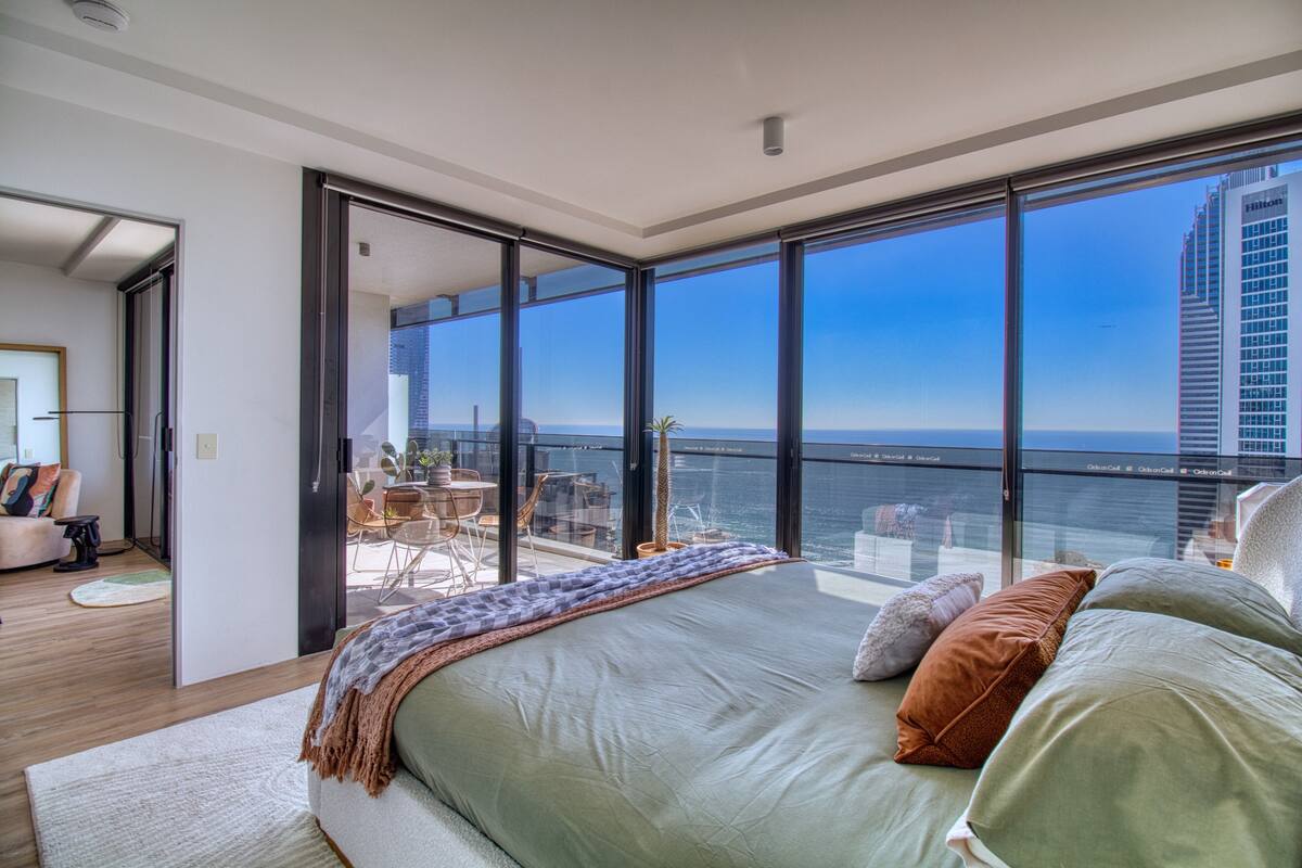A modern bedroom with a large bed, a balcony with a view of the ocean, and a city skyline in the background.