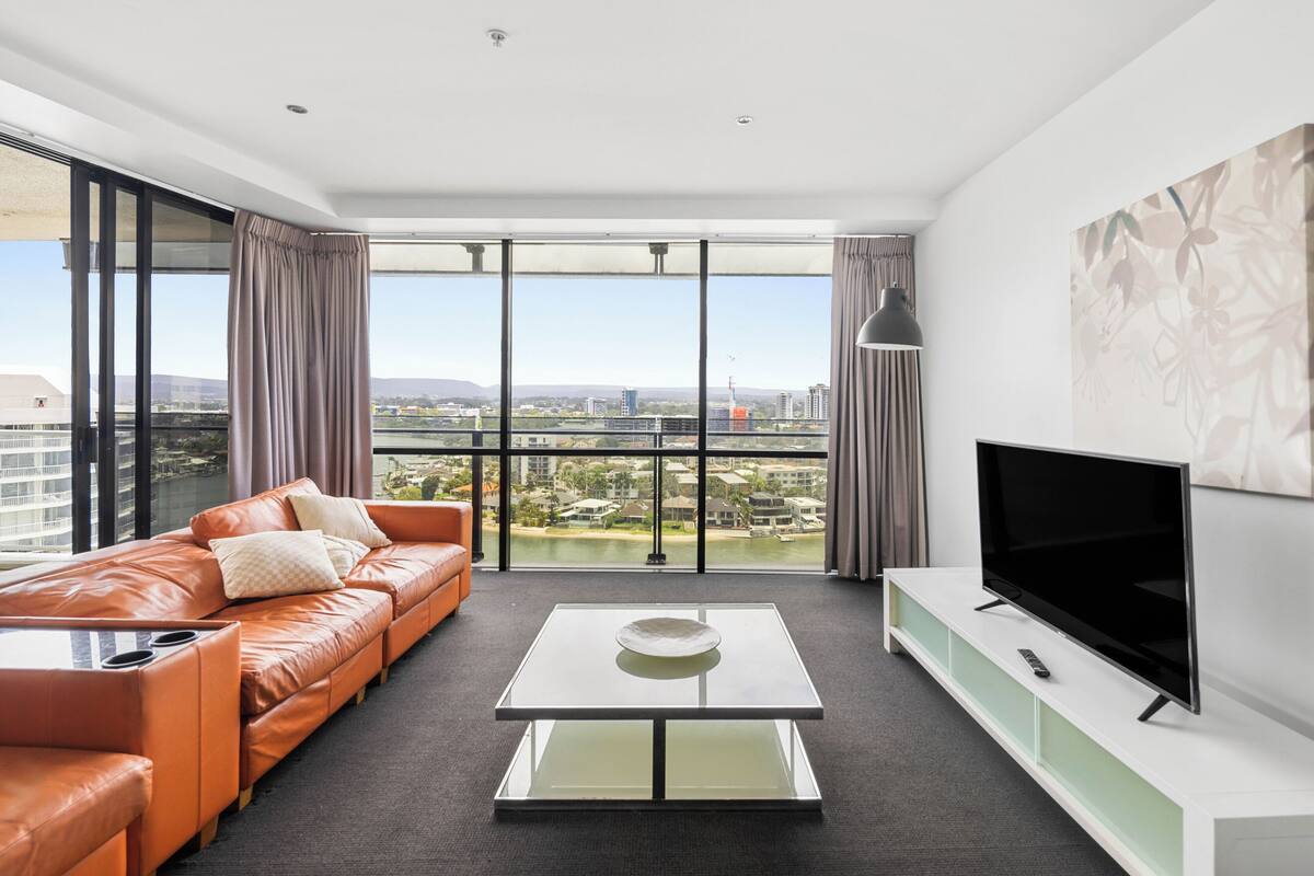 A modern living room with a leather sofa, a glass coffee table, a flat-screen TV, and a city view through large windows.