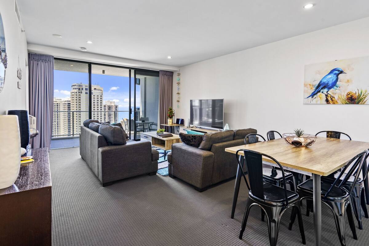 A modern living room with a balcony, grey sofas, a wooden dining table, and a flat-screen TV.
