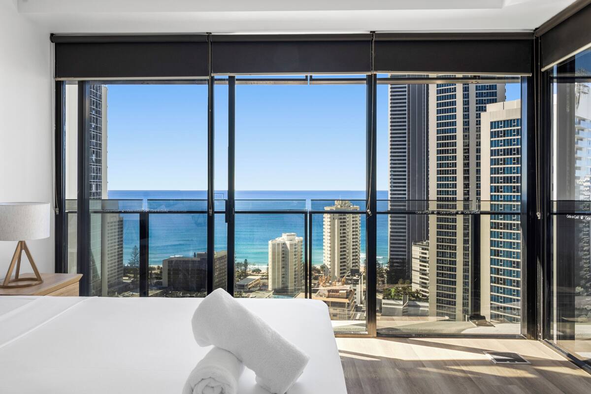 A hotel room with a large window offering a view of the ocean and city skyline.