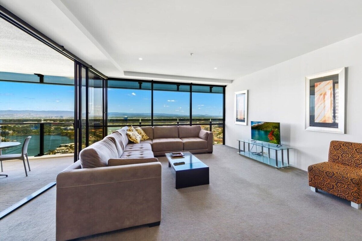 A spacious living room with a large sofa set, a coffee table, and a TV on a glass stand. The room has large windows offering a view of the landscape.