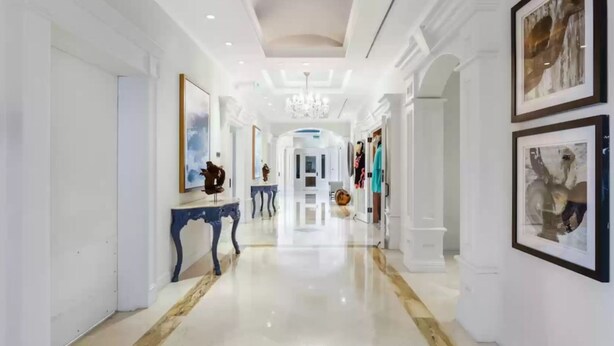 A spacious, well-lit hallway with marble flooring, framed artwork on the walls, and a central chandelier.