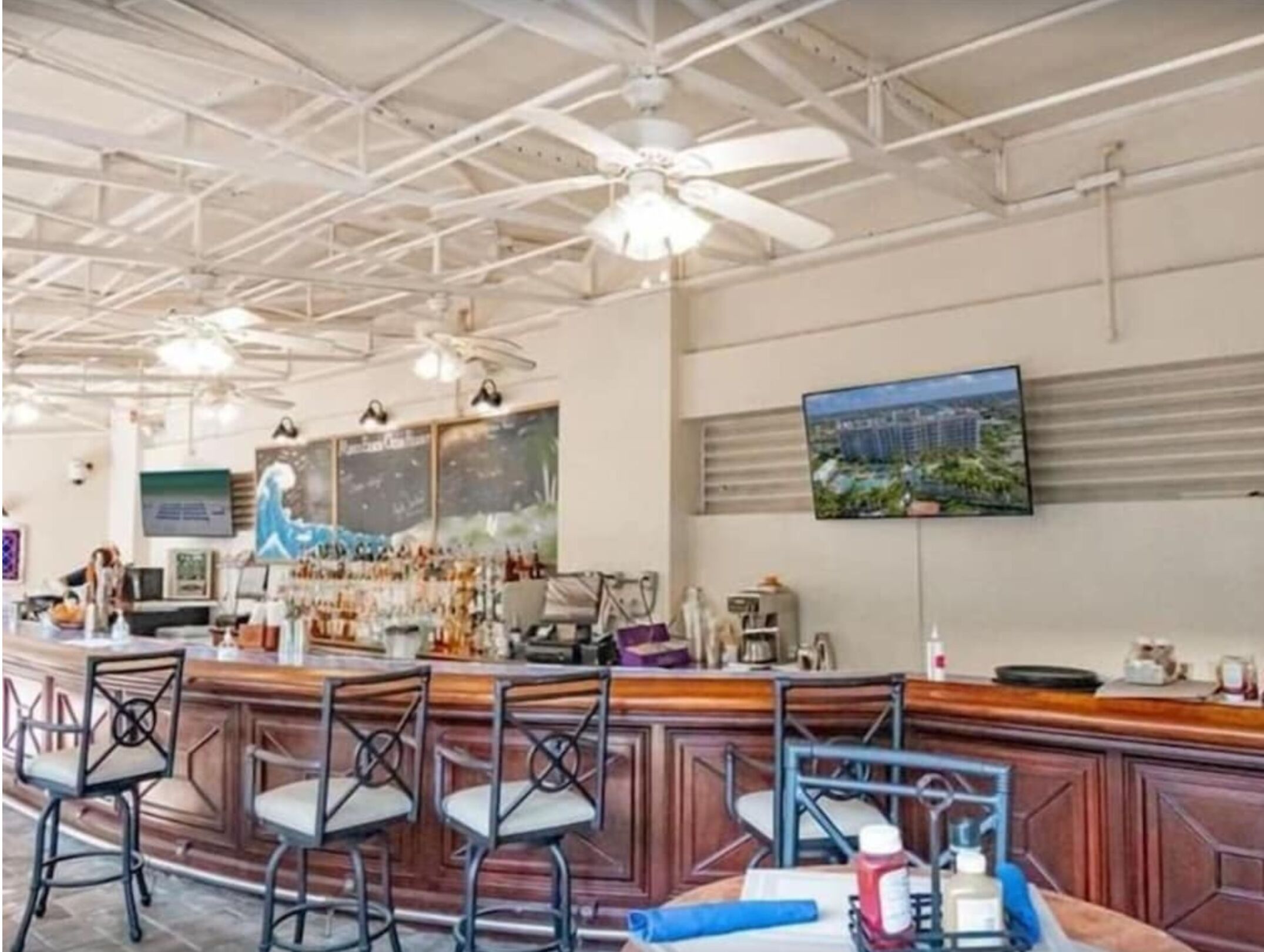 Uma área de bar com balcão de madeira e cadeiras de metal, ventiladores de teto e uma televisão fixada na parede.