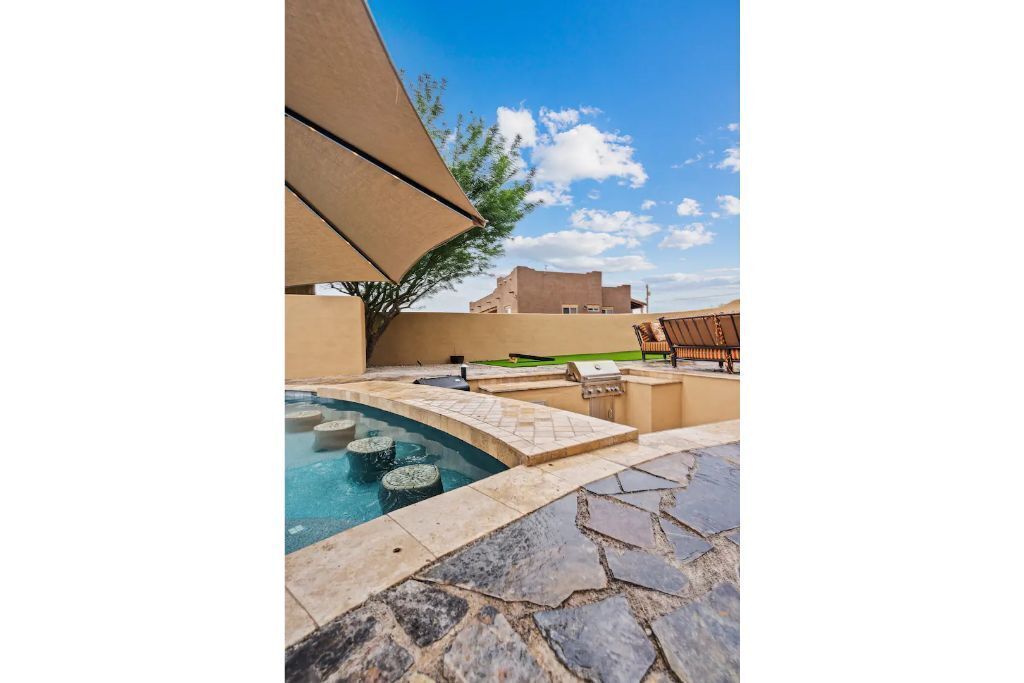 Zona de piscina al aire libre con baldosas de piedra, una parrilla y una sombrilla grande que da sombra.