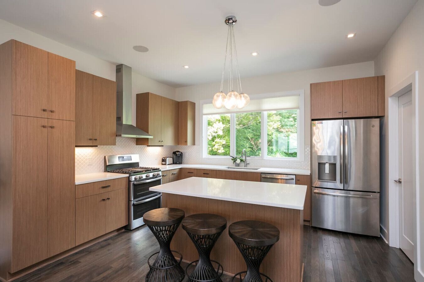 A modern kitchen with wooden cabinets, a stainless steel refrigerator, a central island with stools, and a window providing a view of greenery.