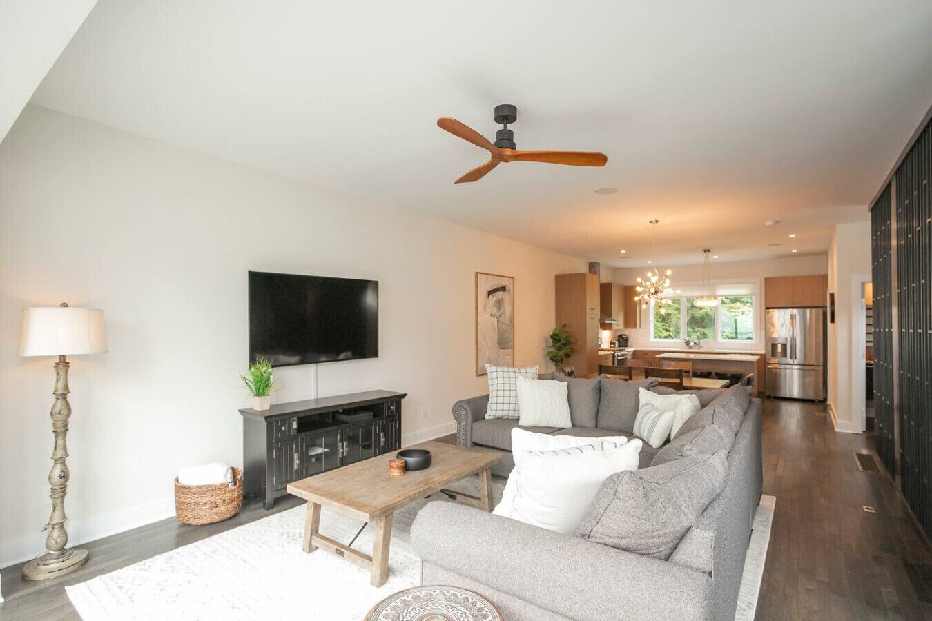 A modern living room with a large sofa, a coffee table, and a flat-screen TV. There is a ceiling fan and a kitchen in the background.