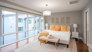 A bedroom with a large window, a bed with white and orange bedding, a bedside table with a lamp, and a small bench with towels.