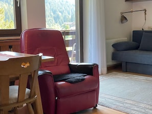Un soggiorno con una poltrona in pelle rossa, un divano blu, un tavolo in legno e vista su una foresta attraverso una finestra.