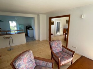 Ein Zimmer mit zwei gemusterten Sesseln, Holzboden, einer Küchenzeile mit Barhocker und Blick durch eine offene Tür.