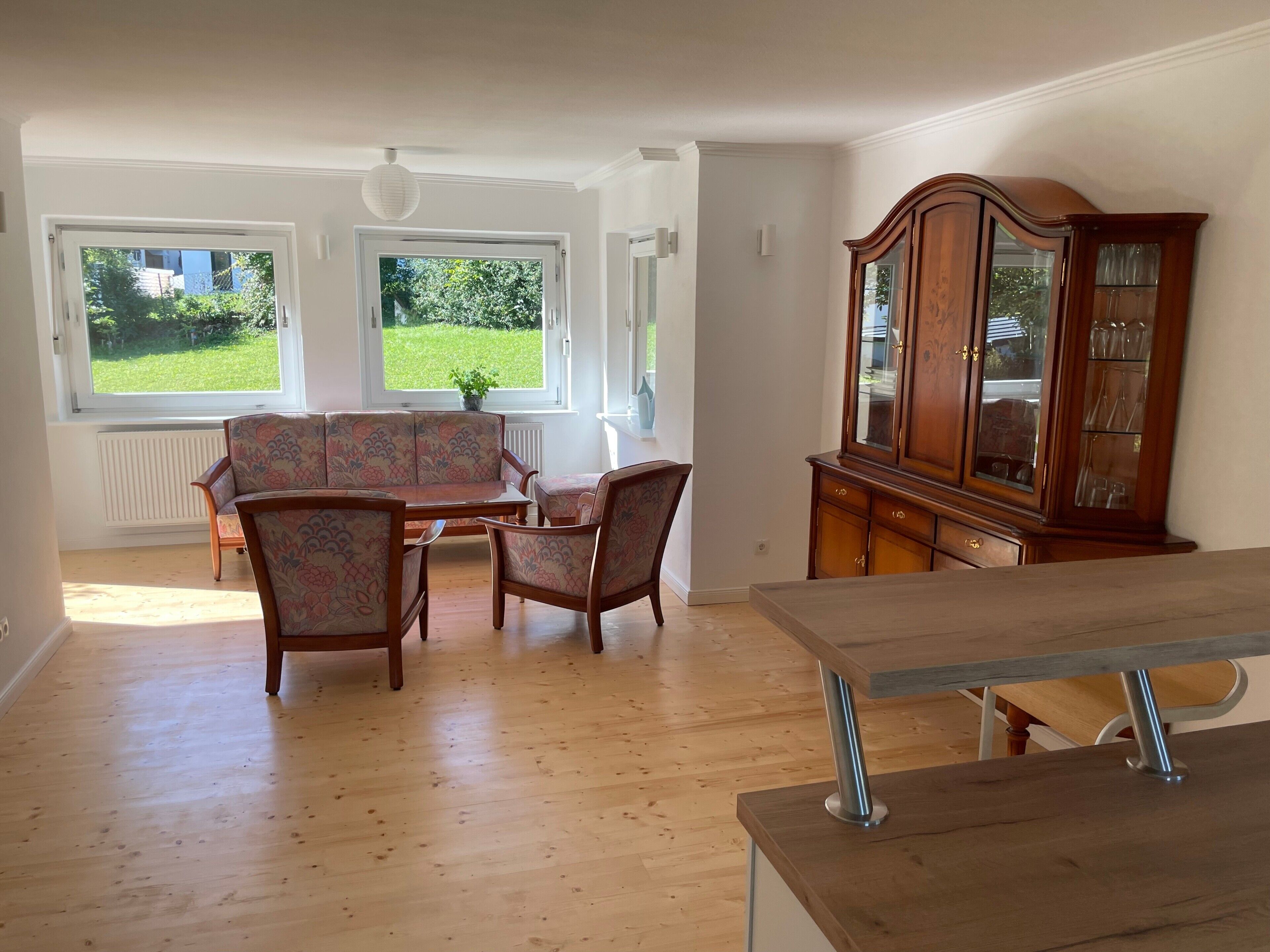 Um quarto com piso de madeira, uma mesa de jantar com cadeiras, um armário com portas de vidro e duas janelas com vista para o verde.
