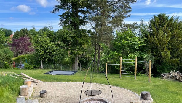 A circular fire pit surrounded by wooden stumps, a trampoline, and a wooden swing set in a grassy backyard. - Holiday Home "Hofberg Villa" with Terrace, Open Garden and High-speed Wi-Fi (Landshut)