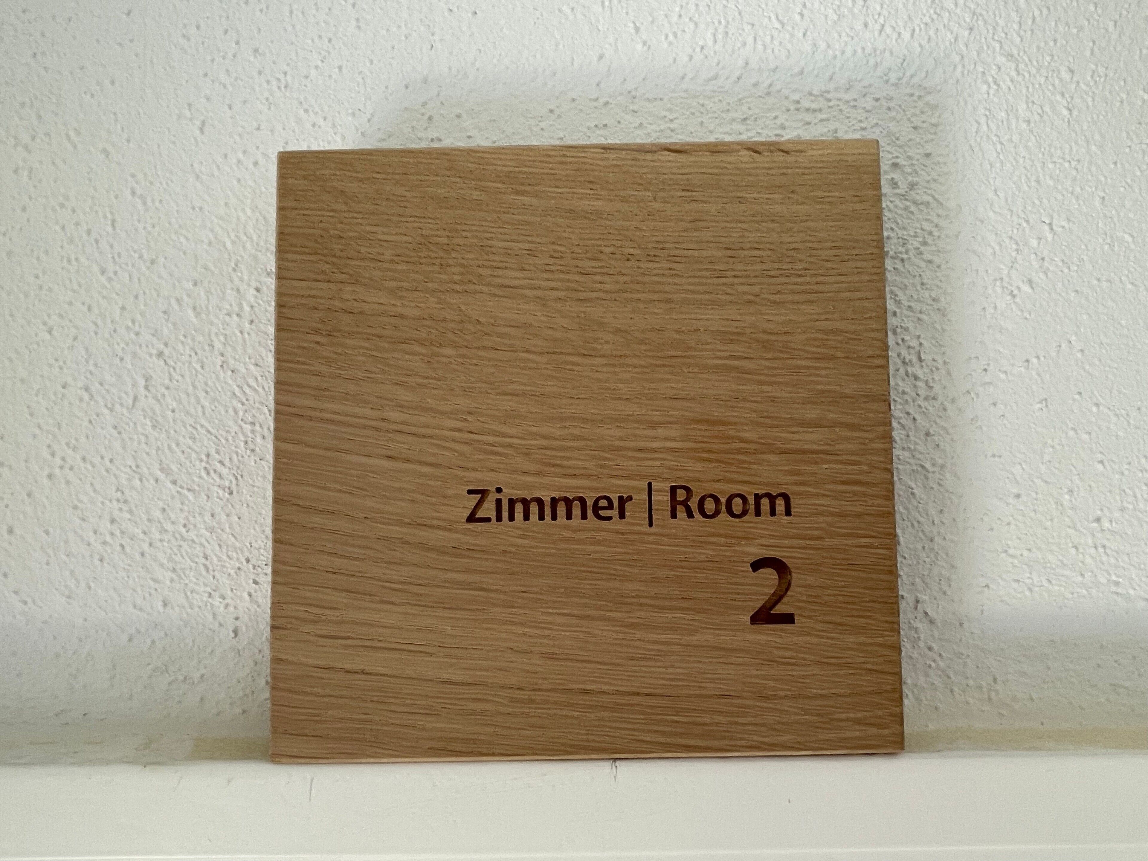 Ein Holzschild mit der Aufschrift 'Zimmer | Room 2', das eine Zimmernummer angibt.