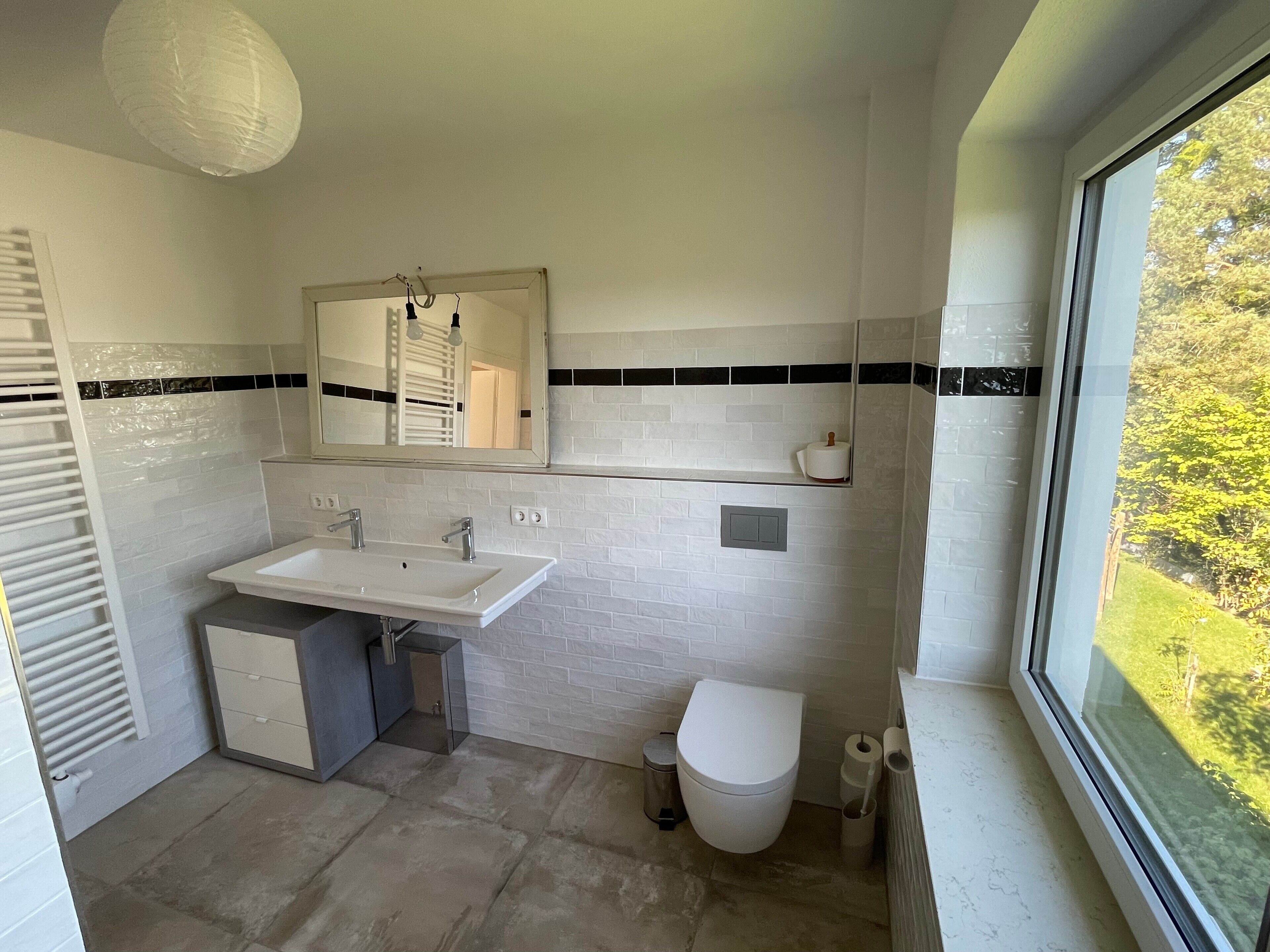 Een badkamer met een witte wastafel, spiegel en toilet, een raam met uitzicht op groen, en een handdoekhouder.
