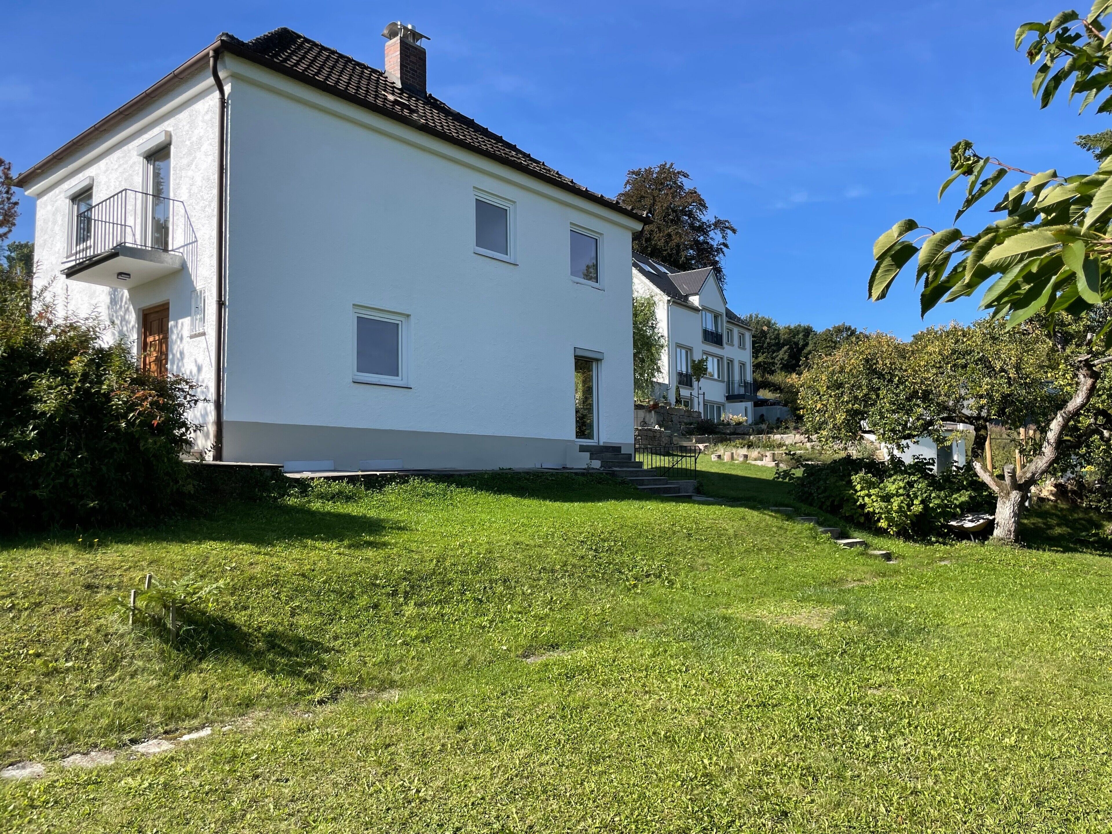 Une maison blanche à deux étages, dotée d’un balcon, d’une cheminée et d’une pelouse bien entretenue.