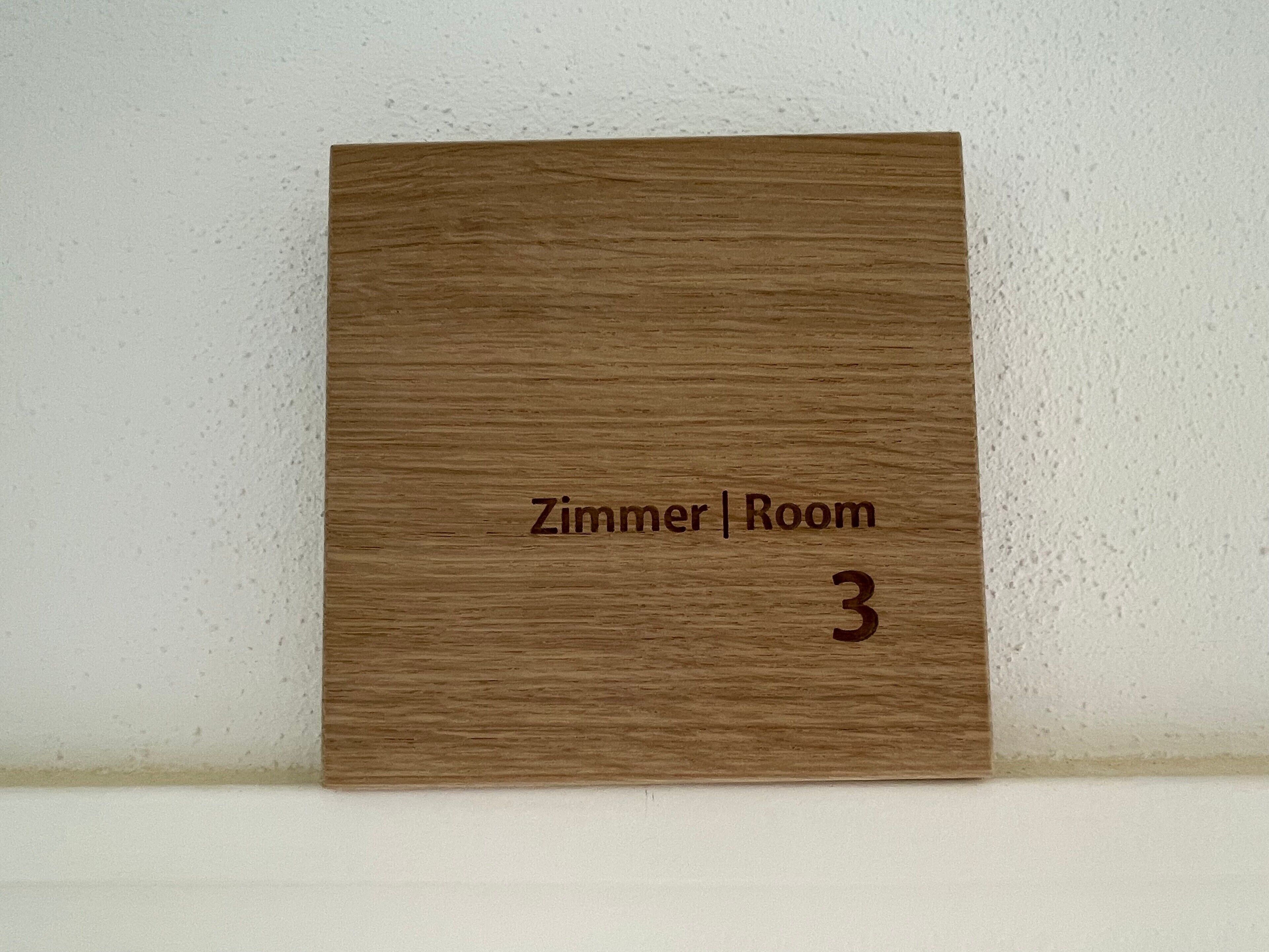 Un letrero de madera con la inscripción 'Zimmer | Room 3' que indica un número de habitación.