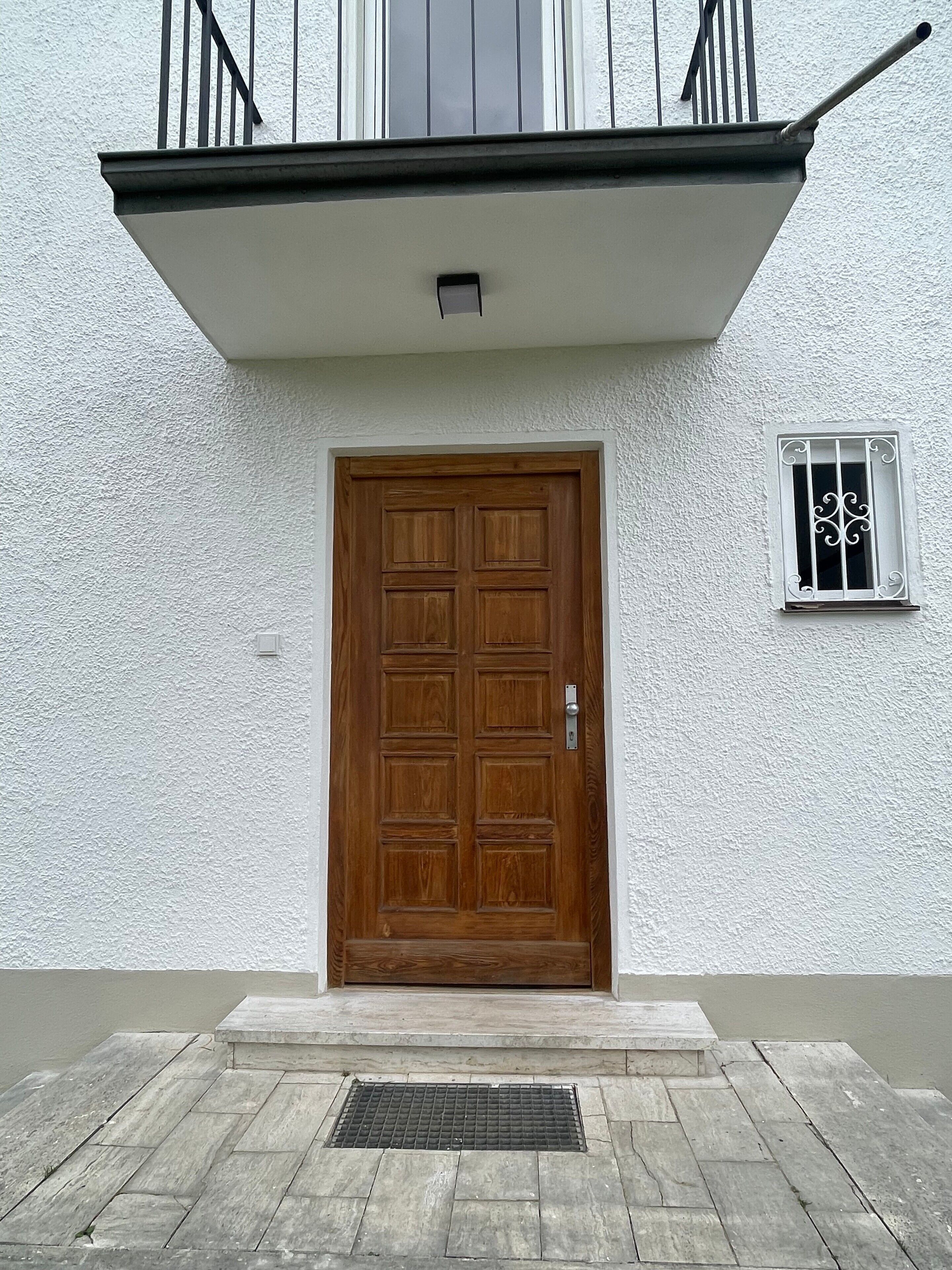 A wooden door with a metal handle, a small window with decorative bars, and a metal grate on the ground.