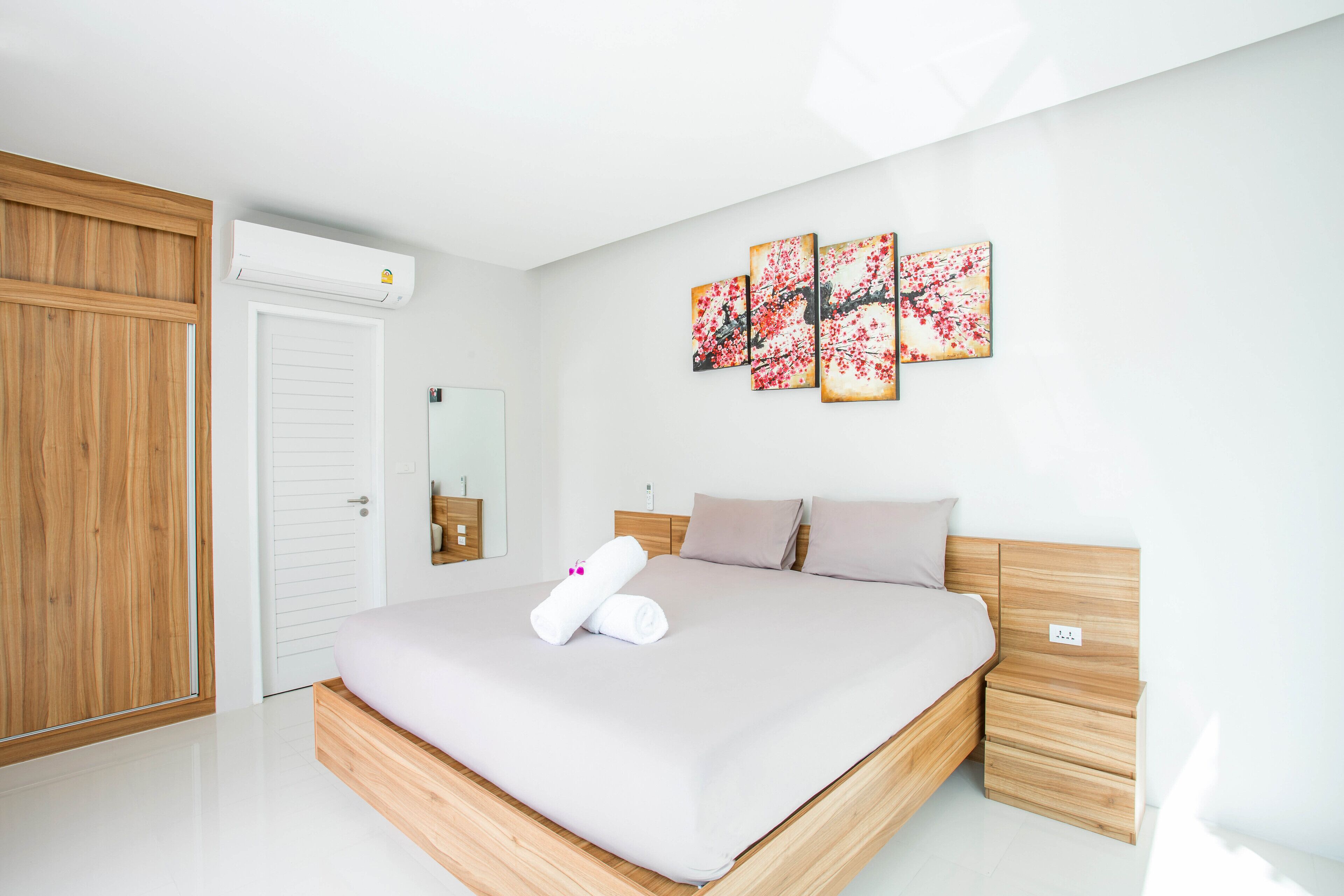A modern bedroom with a wooden bed frame, a white mattress, and a wooden nightstand. There is a wall-mounted air conditioning unit and a set of three framed artworks above the bed.