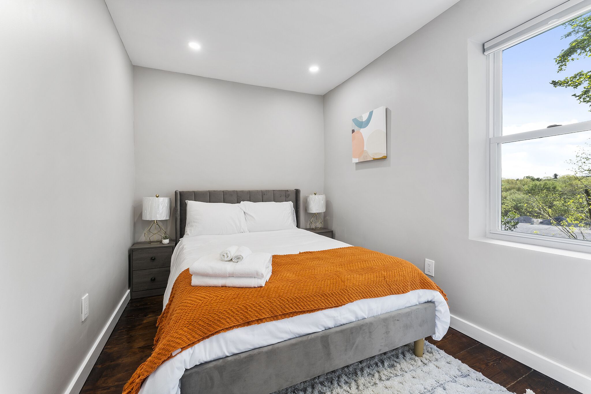 A modern bedroom with a bed, nightstands, a window, and a view of greenery outside.