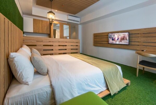 Een moderne slaapkamer met een houten hoofdbord, een bed met wit beddengoed, een groene poef, een wandgemonteerde televisie en een plafondlamp.