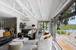 Een moderne woonkamer met een zwarte leren bank, witte fauteuils en een salontafel. Een groot raam biedt uitzicht op een meer en groen.