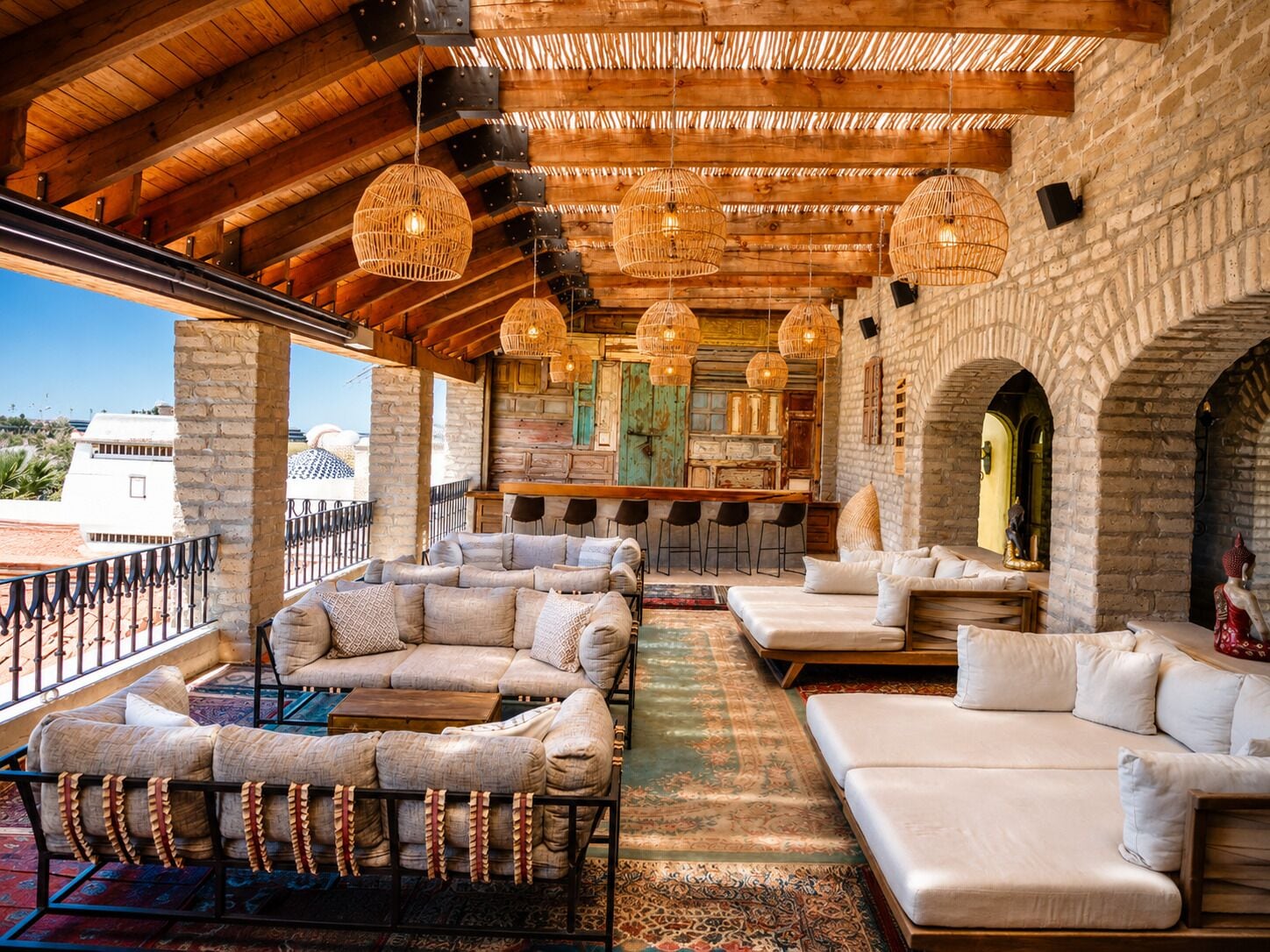 A spacious outdoor seating area with a wooden ceiling, wicker pendant lights, and a stone wall backdrop.