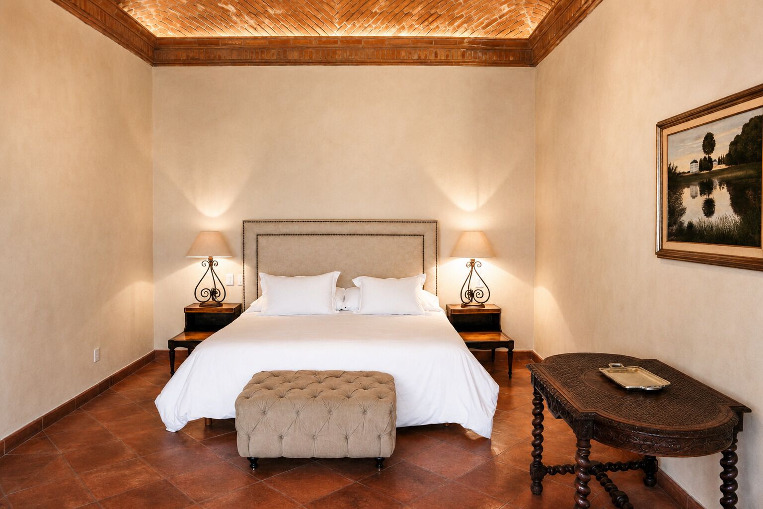 A bedroom with a large bed, two bedside tables with lamps, a small ottoman, and a framed picture on the wall.