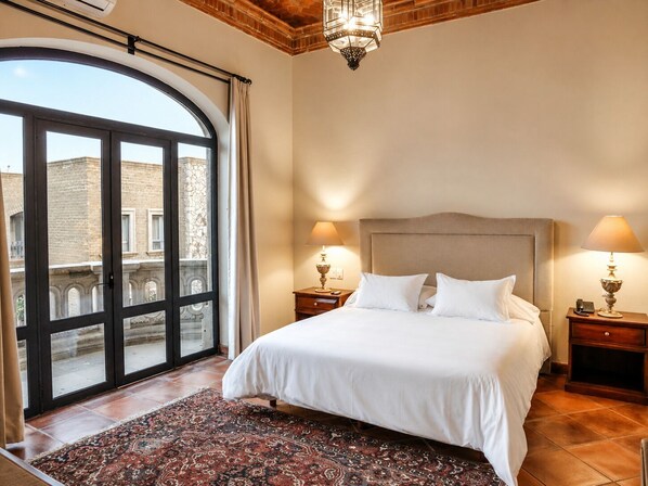 Une chambre à coucher avec un grand lit, deux tables de chevet, un lustre et une vue sur l’extérieur, visible à travers une porte vitrée.