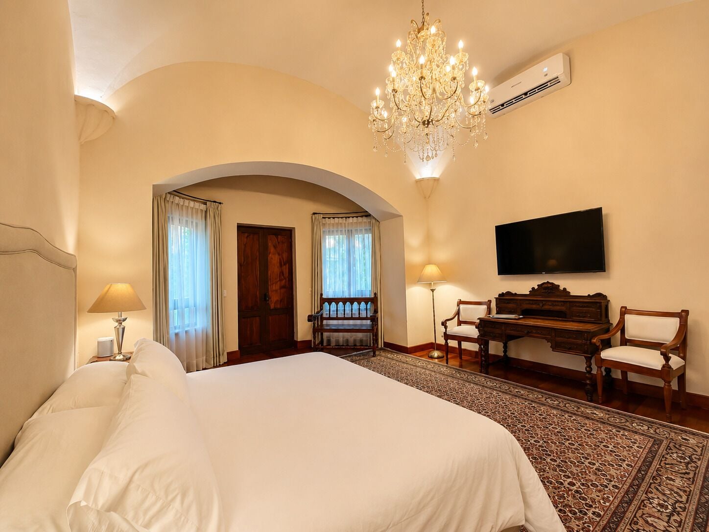 A hotel room with a large bed, a chandelier, a television, a desk, and a patterned rug.