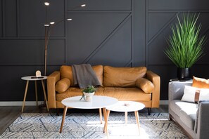 Uma sala de estar moderna com um sofá de couro marrom, duas mesas redondas brancas, um abajur alto e uma grande planta verde.