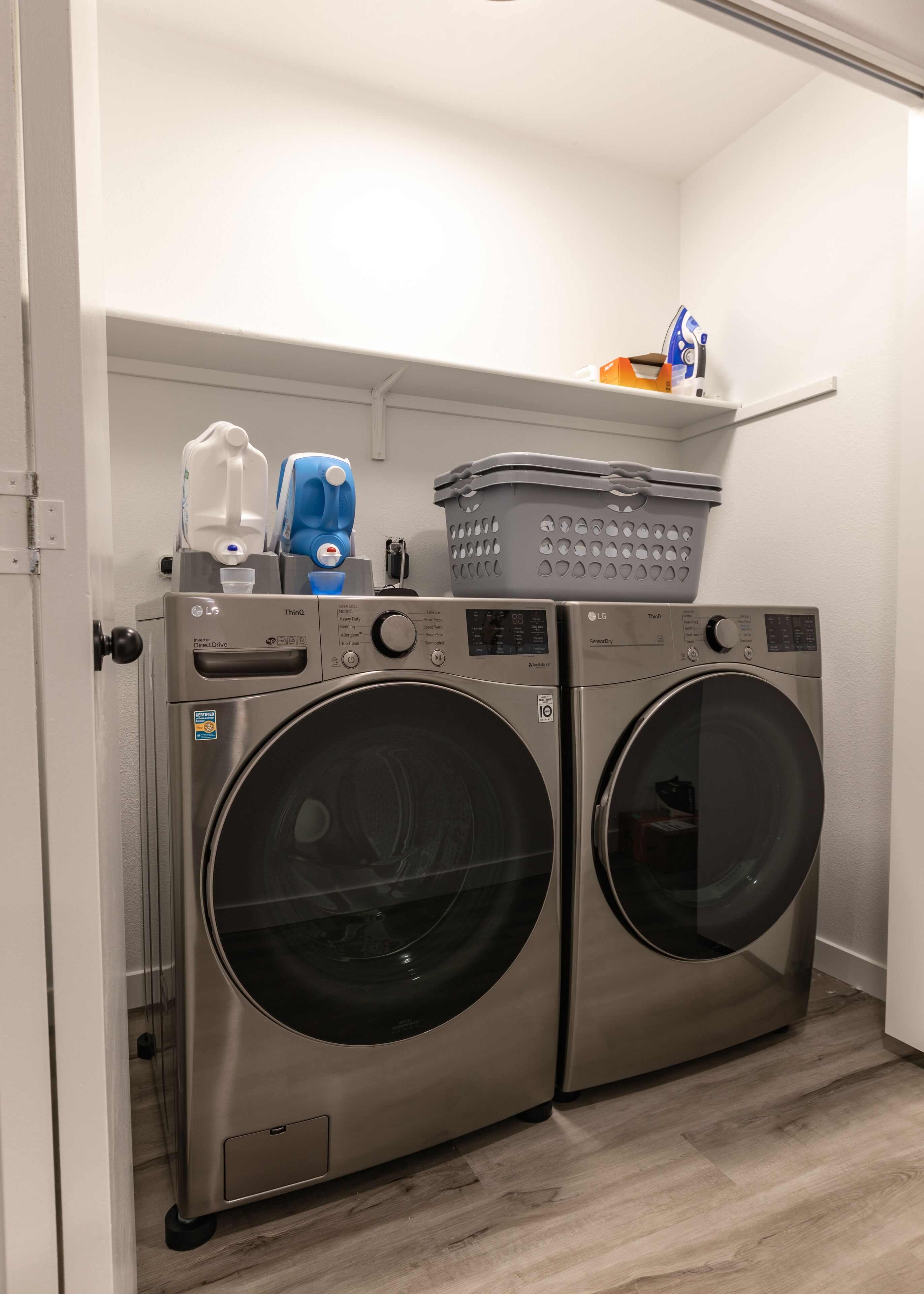 A laundry room with two front-loading washing machines, a drying rack, and a shelf with laundry supplies.